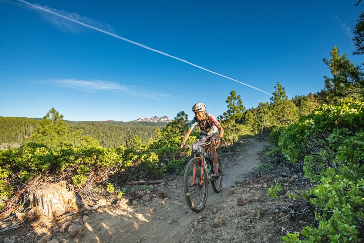 a beautiful high altitude shot in Oregon of a rider on a single track, with the distant forest and far off mountains below and in the background 